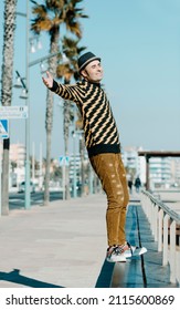 A Smiley Middle-aged Man, Wearing Hat And Modern Casual Clothes, Is About To Fall Backwards From The Bench Where Is Standing In The Promenade Next To A Urban Beach