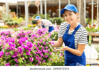 Smilign enthusiastic young female employee of garden center in striped t-shirt and blue dungarees offering lush blooming magenta bougainvillea - Powered by Shutterstock