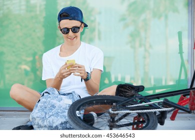 Smiling 15-year old schoolboy in street wear and stylish shades messaging via social networks with happy look, relaxing in the shadow on sunny day during his ride on BMX bike in the city center - Powered by Shutterstock