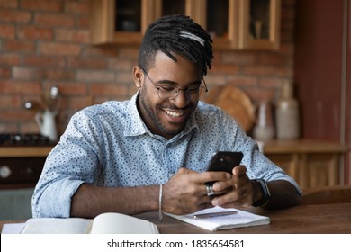 Smiling 20s African American Man Sit At Home Kitchen Office Text Message On Smartphone Online. Happy Young Biracial Male In Glasses Use Cellphone Browse Surf Wireless Internet On Modern Gadget.