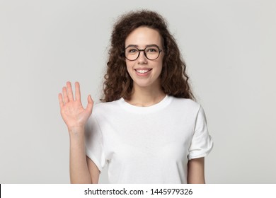 Smiling 20s Girl Wearing Glasses White T-shirt Posing Isolated On Gray Studio Background, Woman Looking At Camera Wave Hand Greeting Get Acquainted With Someone Polite Gesture Nice To Meet You Concept