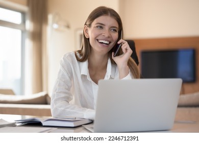 Smiling 25s woman sit at desk with laptop and talks on smartphone, blab with friend, lead formal pleasant conversation, providing services to client using telecommunications mobile operator company - Powered by Shutterstock