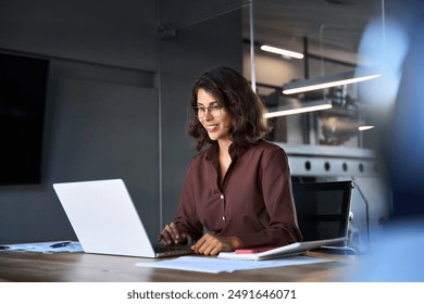 Smiling 30s latin hispanic middle-aged business woman working on laptop computer in modern office. Indian young businesswoman professional employee using pc doing online banking analysing at workplace