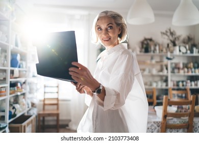 Smiling 50 years old caucasian businesswoman hold clipboard and look at camera at home art studio with shelves full of ceramic products. Small business and entrepreneurship. Modern successful woman