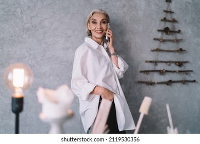 Smiling 50 years old caucasian businesswoman standing at wall, talking on smartphone and looking at camera. Small business and entrepreneurship. Modern successful woman. Art studio with decoration