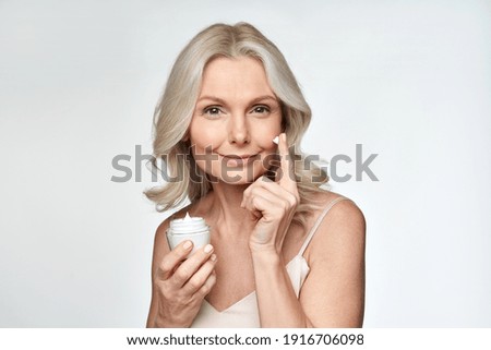 Smiling 50s middle aged mature woman putting tightening facial cream on face looking at camera. Anti age healthy dry skin care beauty therapy concept, skincare rejuvenation treatment against wrinkles.