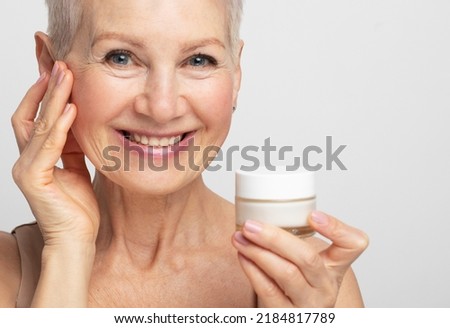 Smiling 60s middle aged mature woman putting tightening facial cream on face looking at camera. Anti age healthy dry skin care beauty therapy concept, skincare rejuvenation treatment against wrinkles.