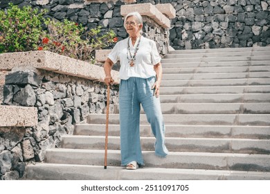 Smiling 70 year old elderly woman with walking disability holding stick knob while walking down stairs outdoors, leaning on stick, suffering from muscle problems, bone disease, recovering from trauma - Powered by Shutterstock