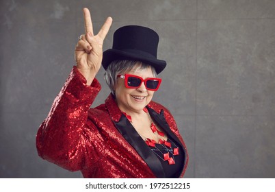 Smiling Active Grandma In The Mood To Party. Happy Positive Funny Retired Senior Woman In Black Top Hat, Cool Glasses, Jewelry And Bling Sequin Concert Outfit Doing V Sign On Gray Studio Background