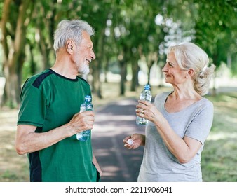 Smiling Active Senior Couple Holding Water Bottles, Drinking And  Jogging Together In The Park