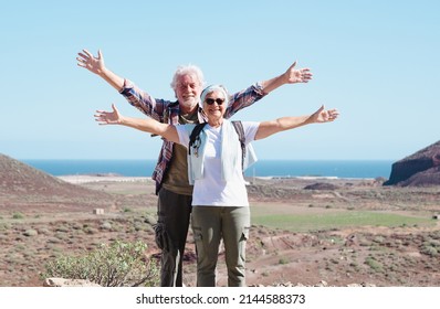 Smiling Active Senior Couple With Outstretched Arms In Outdoors Excursion Hiking In Mountain Enjoying Healthy Lifestyle.  Scenic View Of Sea And Mountain Background