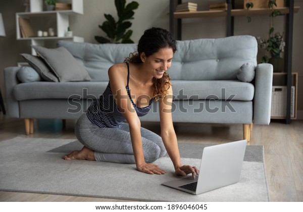 Smiling active young Caucasian woman look at\
laptop screen train at home on online program on computer. Happy\
millennial sporty female practice yoga or pilates in living room\
have webcam lesson.