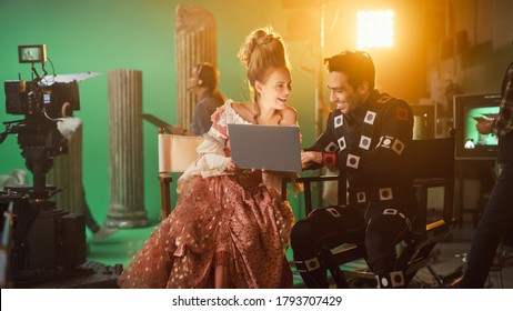 Smiling Actress Wearing Renaissance Dress And Actor Wearing Motion Capture Suit Sitting On Chairs, Use Laptop Computer. Studio Shooting High Budget Movie. On Film Studio Period Costume Drama Film Set