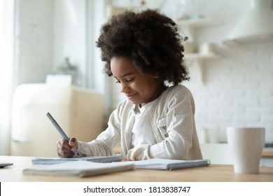 Smiling adorable beautiful african ethnicity little girl sitting at table, writing notes in copybook. Happy small school aged mixed race kid involved in preparing interesting homework alone indoors. - Shutterstock ID 1813838747