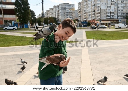 Smiling adorable Caucasian teen boy, expressing positive emotions while feeding rock pigeons and feral doves in the urban square. The concept of people, nature and kindness for animals