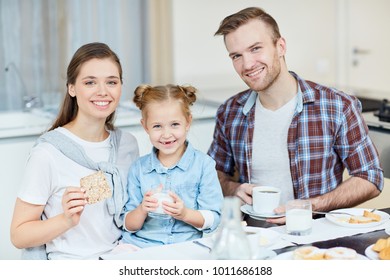 Smiling adorable girl with glass of fresh milk and her parents sitting by table and having breakfast