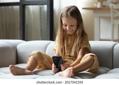 Smiling Adorable Little Girl Using Modern Smartphone Sitting On Cozy Couch, Happy Cute Preschool Child Kid Having Fun With Device, Holding Phone, Playing Game, Watching Funny Video In Social Network