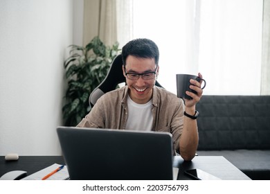 Smiling Adult Asian Man Wearing Glasses Holding Cup Of Tea Or Coffee Using Laptop At Home. Remote Job, Freelance Working Concept.