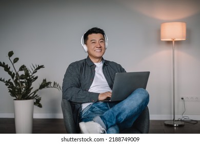 Smiling Adult Asian Man In Wireless Headphones Enjoy Favorite Music, Typing On Laptop, Watch Video, Sit In Armchair In Minimalist Room Interior. Work With Device Alone At Home, Online Call, Business