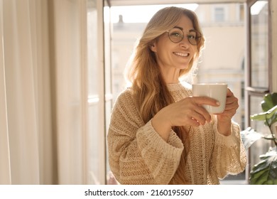 Smiling Adult Caucasian Woman Drinks Tea Or Coffee From Cup Against Background Of City Apartment Window. Blonde Wears Glasses And Sweater. Peace Of Mind And Mental Health.