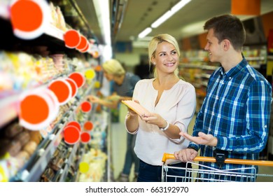 Smiling adult customers with trolley looking at chilled sausages. Focus on the woman