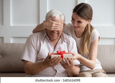 Smiling adult daughter closing older father eyes, preparing surprise, holding gift box with red bow in hand, young woman congratulating dad with birthday or fathers day, sitting on couch at home
