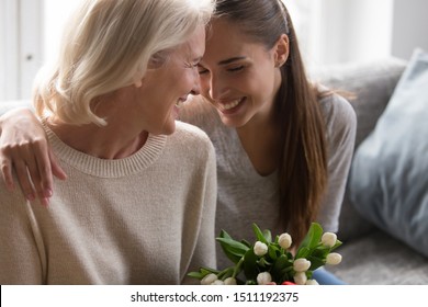 Smiling adult daughter greeting happy senior 50 mother with anniversary presenting bouquet of flowers, happy elderly mom and grownup child hug embrace celebrate birthday at home together