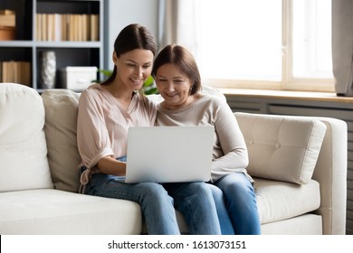 Smiling Adult Daughter And Middle-aged Mother Sit Relax On Couch In Living Room Using Laptop Together, Happy Grownup Girl Child And Senior Mum Rest On Sofa Browse Internet Shopping Online On Computer