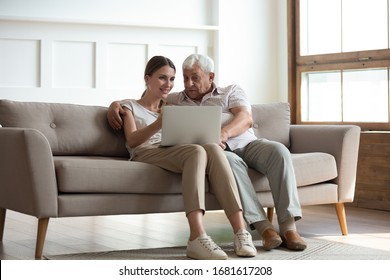 Smiling Adult Daughter Teaching Older Father To Use Computer, Pointing At Screen, Explaining, Happy Young Woman And Mature Dad Or Grandfather Hugging, Using Laptop Together, Sitting On Couch At Home