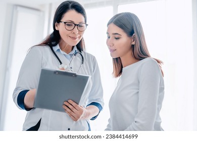 Smiling adult female doctor reporting medical results to young girl patient, explaining diagnosis, showing analysis tests, planning pregnancy. - Powered by Shutterstock