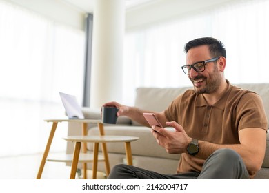 Smiling adult man holding phone browses through social networks while drinking coffee in the comfort of his home. Young entrepreneur working at home taking break to drink nice warm cup of coffee - Powered by Shutterstock