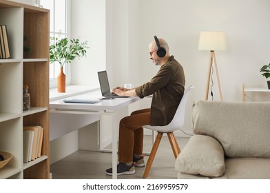 Smiling adult man listening to music on headphones while working on laptop at home. Side view of middle-aged man listening to his favorite music, searching information on Internet or making video call - Shutterstock ID 2169959579