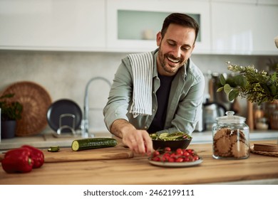 A smiling adult man taking a cherry tomato to mix it with the fresh and healthy vegetable salad he made for breakfast while leaning on the kitchen counter. - Powered by Shutterstock