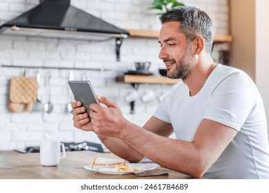 Smiling adult man using holding digital tablet pad wireless device gadget while eating breakfast sandwich at kitchen. Middle age guy looking watching video reading news checking email social media - Powered by Shutterstock