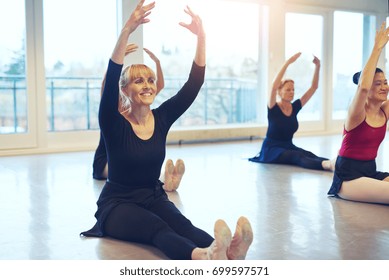 Smiling Adult Women Sitting On Floor With Hands Up While Doing Ballet Gymnastics In The Class.