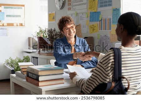 Smiling advisor shaking hands with client while sitting at desk, surrounded by office materials and diagrams. Engaging meeting in office setting