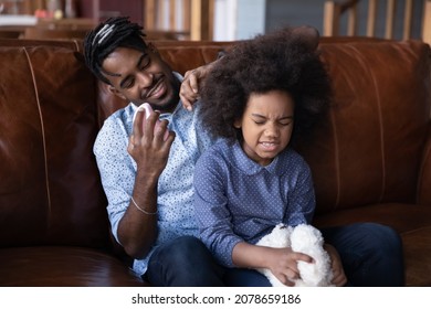 Smiling affectionate African ethnicity young father combing detangling naughty curly hair of small kid daughter, helping with difficult hairstyle, sitting together on comfortable couch in living room. - Powered by Shutterstock