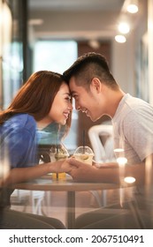 Smiling affectionate young Asian couple holding hands and touching foreheads while dating in cafe