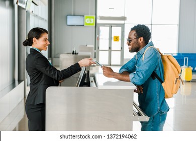 Smiling African American Airport Worker Taking Passport And Air Ticket From Tourist With Backpack