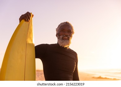 Smiling African American Bearded Senior Man With Surfboard Standing At Beach Against Clear Sky. Sunset, Copy Space, Unaltered, Retirement, Aquatic Sport, Holiday And Active Lifestyle Concept.