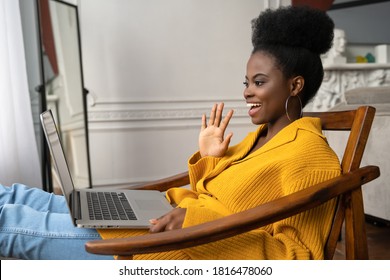 Smiling African American Biracial Millennial Woman With Afro Hairstyle In Yellow Cardigan Resting, Sitting On Armchair, Talking In Video Chat With Her Friends, Say Hello. 