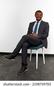 Smiling African American Black Man In Business Suit Sitting On Chair With Crossed Fingers White Wall Background. Full Size Portrait Of Happy Black Man Chief Executive Officer Waiting For Interview