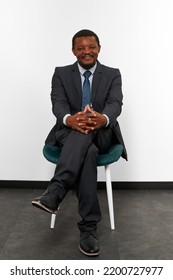 Smiling African American Black Man In Business Suit Sitting On Chair With Crossed Fingers White Wall Background. Full Size Portrait Of Happy Black Man Chief Executive Officer Waiting For Interview