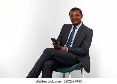 Smiling African American Black Man In Business Suit Sitting On Chair With Smartphone, White Wall Background. Happy Black Businessman, Chief Executive Officer Satisfied Successful Interview