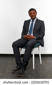 Smiling African American Black Man In Business Suit Sitting On Chair With Crossed Fingers White Wall Background. Full Size Portrait Of Happy Black Man Chief Executive Officer Waiting For Interview