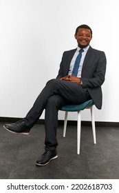 Smiling African American Black Man In Business Suit Sitting On Chair With Crossed Fingers White Wall Background. Full Size Portrait Of Happy Black Man Chief Executive Officer Waiting For Interview