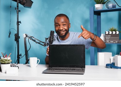 Smiling african american blogger pointing at laptop with black screen while recording product presentation. Vlogger presenting digital device and streaming review on social media - Powered by Shutterstock