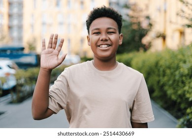 Smiling African American boy with curly hair and brown skin stands on a bustling city street. Smiling and waving cheerfully on a sunny day - Powered by Shutterstock