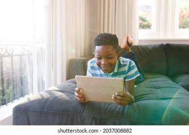 Smiling african american boy using tablet and lying on couch in living room. spending time alone with technology at home. - Powered by Shutterstock