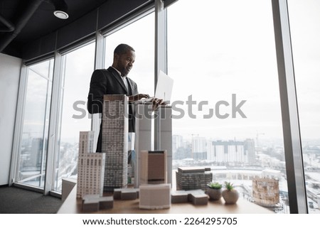 Smiling african american business man real estate agents in black stylish formal suit standing with laptopnear 3d model skyscrapers of city architecture design at office with panoramic city view. [[stock_photo]] © 